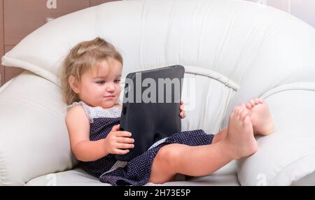 La bambina si trova sulla sedia e gioca sul tablet Foto Stock