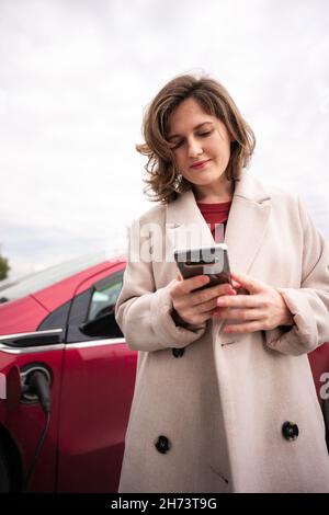 Donna in cappotto beige con smartphone accanto all'auto elettrica rossa Foto Stock