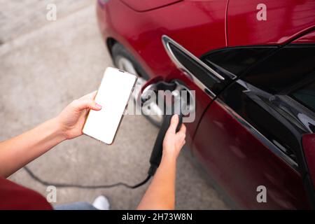 Mani con smartphone e spina di ricarica sullo sfondo dell'auto elettrica . Schermo bianco, puoi aggiungere i tuoi contenuti qui Foto Stock