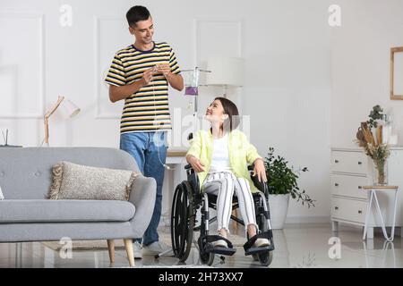 Giovane uomo che mette sua moglie in sedia a rotelle su un gocciolamento a casa Foto Stock