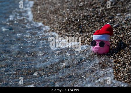 Piggy banca in un cappello di babbo natale e occhiali da sole su una spiaggia di ciottoli vicino al mare. Foto Stock