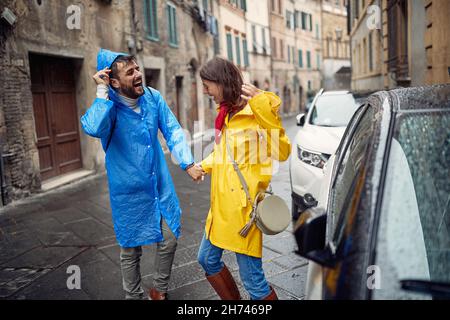 Una giovane coppia allegra in impermeabile sta avendo un buon tempo mentre si sta allontanando dalla pioggia in una giornata nuvolosa in città. Passeggiata, pioggia, città, relationshi Foto Stock