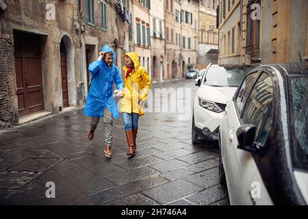 Una giovane coppia allegra in impermeabile si diverte mentre corre dalla pioggia verso l'auto durante una passeggiata in città in una giornata nuvolosa. Cammina, pioggia, cit Foto Stock