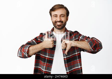Vantaggi e svantaggi. Sorridente ragazzo barbuto mostra pollici su e pollici giù, in piedi su sfondo bianco Foto Stock