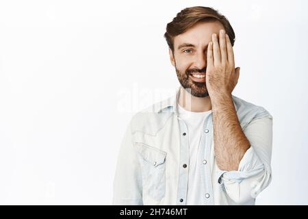 Ritratto di uomo caucasico bearded copre metà del volto e sorride felice alla macchina fotografica, si leva su sfondo bianco Foto Stock