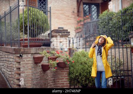 Una giovane ragazza in una strada a piedi in una giornata nuvolosa si nasconde con un impermeabile perché ha iniziato a piovere. Passeggiata, pioggia, città Foto Stock