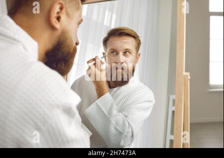 Divertente uomo adulto bearded al mattino applica spazzola alla maschera o crema idratante viso. Foto Stock