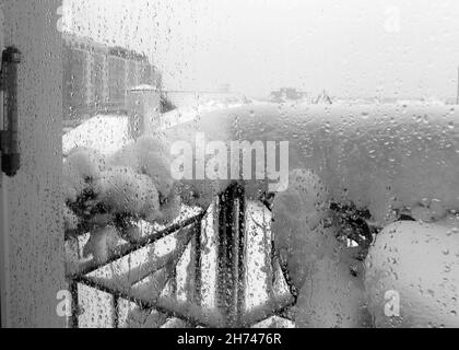 Vista attraverso la finestra sul balcone con tavolo e sedie innevate in inverno. Concentrarsi sulle gocce d'acqua, il termometro mostra una temperatura positiva. Natura Foto Stock