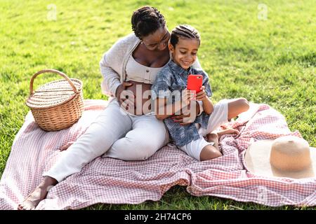 Felice madre incinta africana passare il tempo con suo figlio fare un picnic durante il fine settimana in parco pubblico - la famiglia afro guardare sullo smartphone Foto Stock