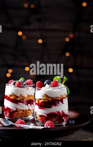 Su sfondo ligneo con luci, dessert con crema, formaggio casolare, lamponi e mirtilli in un bicchiere Foto Stock