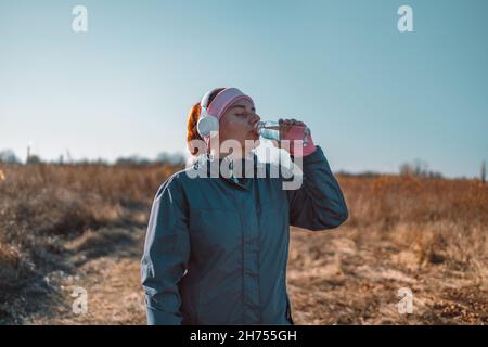 Donna sportiva attiva in abbigliamento sportivo e cuffie wireless bevande acqua fresca da una bottiglia in autunno naturale parco banner sfondo Foto Stock
