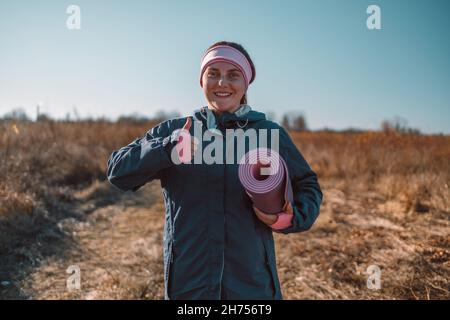 Yoga donna mostra come simbolo con mano tiene materassino di esercizio sembra felice prima di iniziare un allenamento in un parco autunnale. Foto Stock