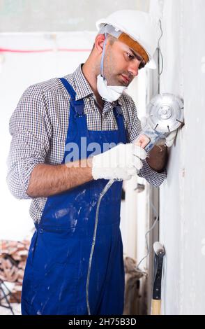 Il riparatore nel casco sta segando la parete con sega circolare Foto Stock