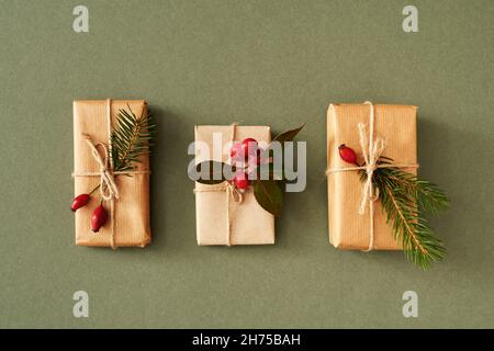 Tre regali di Natale avvolti in carta riciclata con wintergreen, fianchi di rose e rami di abete rosso su sfondo verde Foto Stock