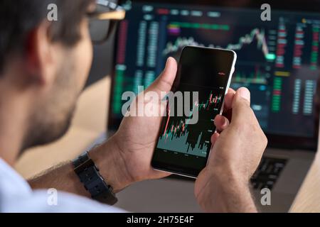 Broker trader tramite telefono che analizza i dati delle tabelle di negoziazione del mercato azionario utilizzando app. Foto Stock