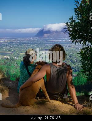 Vista posteriore verticale di una coppia e il loro cane seduto in montagna e ammirando la vista Foto Stock