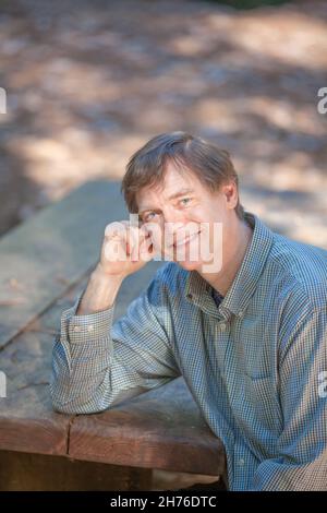 Sorridente uomo caucasico all'inizio degli anni cinquanta seduto da solo al tavolo da picnic in legno nella foresta Foto Stock