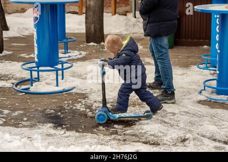 Un ragazzino guida uno scooter accanto al padre su un marciapiede innevato in primavera. Foto Stock