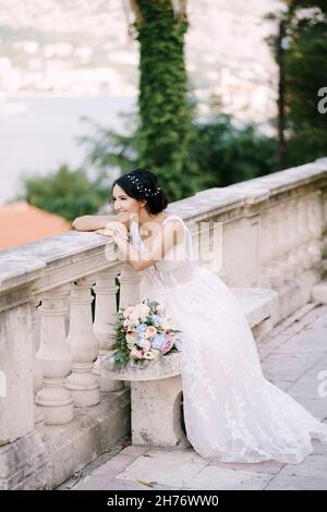 La sposa si siede su una panchina su un sito panoramico nella città vecchia, si appoggia sulla ringhiera e sorride Foto Stock
