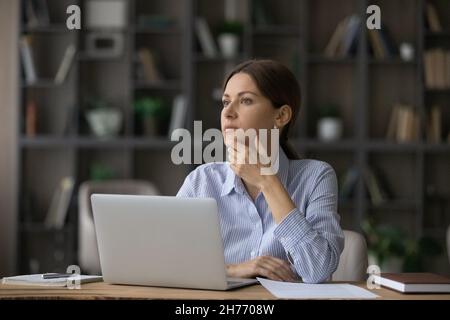 Donna attraente in profondità nel pensiero si siede sul posto di lavoro vicino al laptop Foto Stock