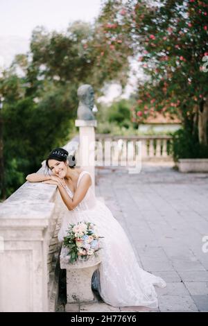 La sposa si siede su una panchina su un sito panoramico nella città vecchia, si appoggia sulla ringhiera e sorride Foto Stock