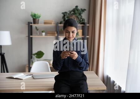 Felice giovane donna d'affari indiana utilizzando cellulare appoggiata sul tavolo. Foto Stock