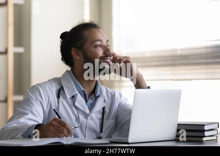 Un uomo medico afro-americano che sorride pensivo prendendo appunti, usando un computer portatile Foto Stock