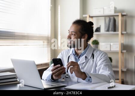 Sorridente medico afroamericano con smartphone, seduto alla scrivania Foto Stock