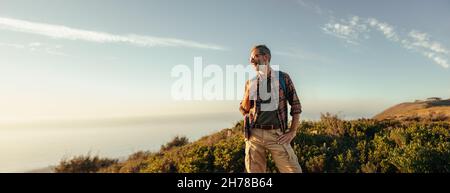 Escursionista che gode della vista panoramica da una collina. Happy escursionista maturo guardando via allegro mentre in piedi sulla cima di una collina con uno zaino. Avventuroso Foto Stock