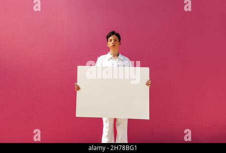 Giovane attivista queer che tiene un banner bianco su sfondo rosa. Ragazzo assertivo che mostra un cartello vuoto in uno studio. Giovane ragazzo gay com Foto Stock