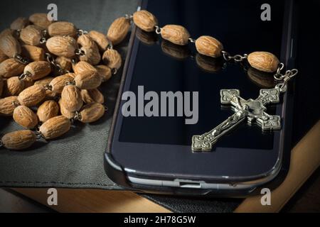 Primo piano estremo di un vecchio crocifisso d'argento con Gesù Cristo e cordone di rosario in legno su uno smartphone nero e la Sacra Bibbia Foto Stock