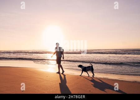 Silhouette di giovane uomo durante la passeggiata con il cane sulla spiaggia di sabbia al bellissimo sorgere del sole. Copia spazio su cielo. Foto Stock