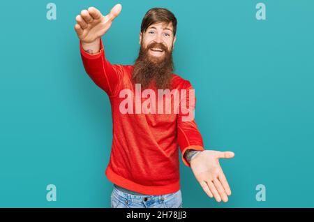 Uomo rosso con barba lunga e vestiti casual guardando la macchina fotografica sorridente a braccia aperte per abbraccio. Espressione allegra che abbraccia la felicità. Foto Stock