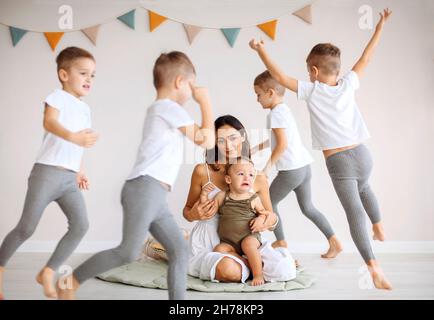 Bambini gemelli che girano intorno alla madre giovane con il bambino piangente, la famiglia grande della mamma e cinque ragazzi bambini che giocano e si divertono insieme a casa. Ragazzo in corsa Foto Stock