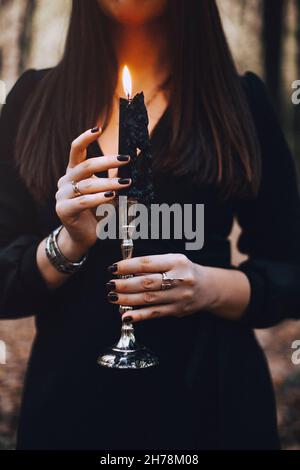 Scatto corto di donna misteriosa in abito nero che tiene la candela sul candelabro nelle sue mani che esegue rituale magico nella foresta d'autunno scuro. Stregoneria c Foto Stock