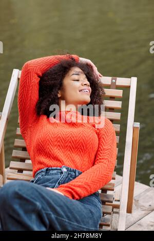 Giovane allegra donna afroamericana rilassata con capelli ricci in arancione caldo maglione rilassante riposarsi sulla poltrona vicino stagno o fiume in autunno weeken Foto Stock