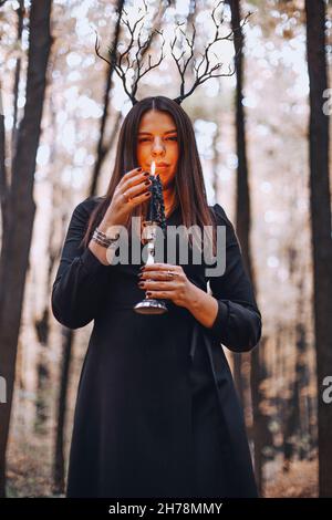 Scatto di donna misteriosa in abito nero che tiene candela su candelabro nelle sue mani che esegue rituale magico nella foresta scura autunno. Concetto di stregoneria Foto Stock