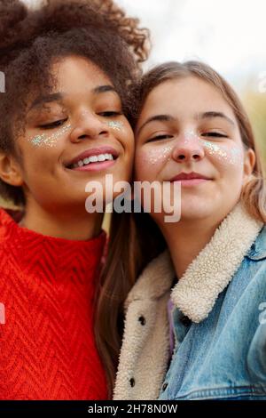 Ritratto di due allegre fidanzate multirazziale sorridenti alla macchina fotografica mentre trascorrono del tempo nella natura autunnale, allegre ragazze teen di razze diverse godendo wa Foto Stock