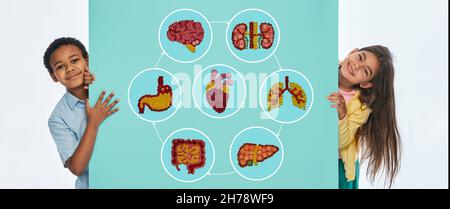 Concetto di salute dei bambini, pediatria. Bambini multietnici vicino alla parete turchese con icone anatomiche con illustrazioni degli organi interni Foto Stock