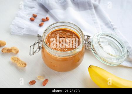 Burro di arachidi in vaso di vetro, arachidi sparse e banana su tavola di legno bianco. Vista dall'alto. Foto Stock