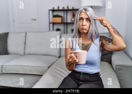 Donna di mezza età dai capelli grigi che beve caffè seduto sul divano a casa sparando e uccidendo se stessi puntando la mano e le dita alla testa come pistola, suic Foto Stock