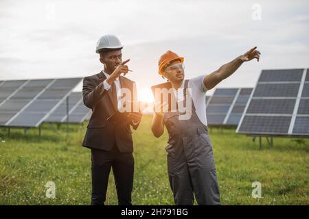 Ingegnere indiano in casco arancione e occhiali di sicurezza che tiene tablet digitale e parlare con l'uomo afro-americano in tuta circa pianta solare, puntando su qualcosa di lontano Foto Stock