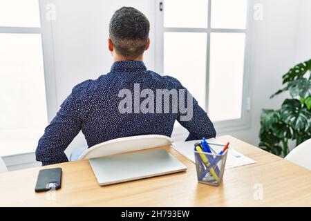 Giovane ispanico con barba che lavora in ufficio con un computer portatile in piedi all'indietro guardando le braccia sul corpo Foto Stock