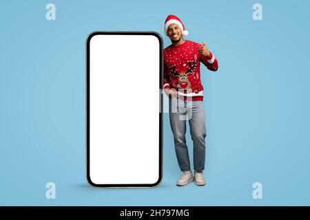 Bel ragazzo nero sorridente in rosso Santa cappello e divertente maglione con cervi in posa con grande cellulare con schermo vuoto e mostrando pollice su, blu studio Foto Stock