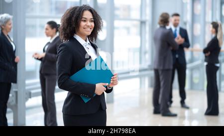 Primo piano ritratto di giovane donna da corsa mista in piedi con cartella documenti di fronte ai colleghi in business building Foto Stock
