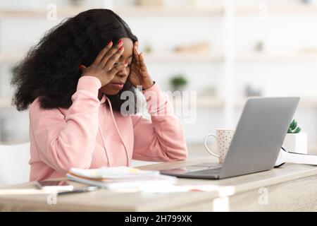Donna nera stressata seduta davanti al laptop, toccando la testa Foto Stock