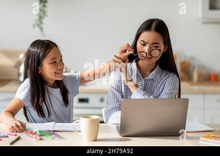 Donna asiatica che lavora su un computer portatile da casa, mentre figlia che la distrava, togliendo gli occhiali della mamma Foto Stock