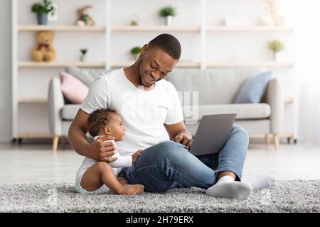 Felice giovane ragazzo nero utilizzando il portatile e babysitter suo bambino a casa Foto Stock