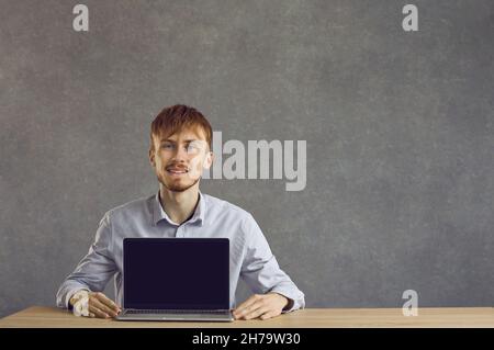 Giovane uomo sorridente seduto al tavolo che mostra un computer portatile con uno schermo vuoto. Foto Stock