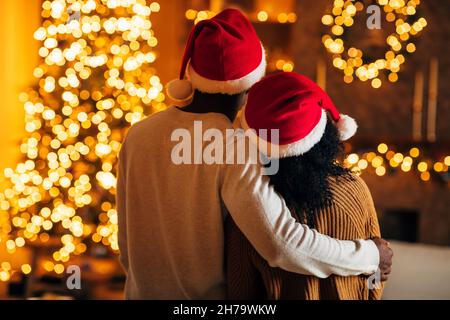 Vista posteriore di giovane coppia nera loving in Babbo Natale cappelli e maglioni hugging, guardando luminoso decorato albero di Natale Foto Stock